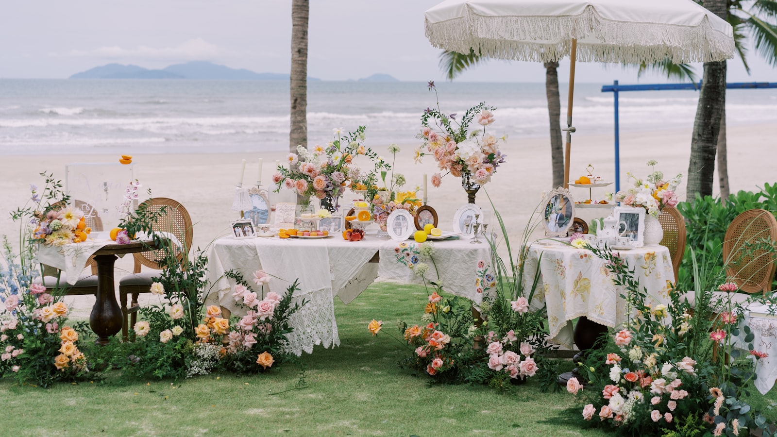 Una mesa de boda vestida en la playa, con flores en tonos coral y crema, una sombrilla de flecos, palmeras y el mar al fondo
