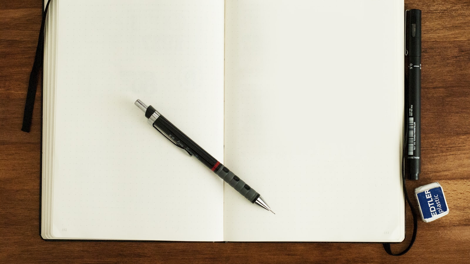 Un cuaderno de tapa dura abierto con las páginas en blanco, un portaminas apoyado encima y más bolígrafos y una goma de borrar al lado, sobre una mesa de madera