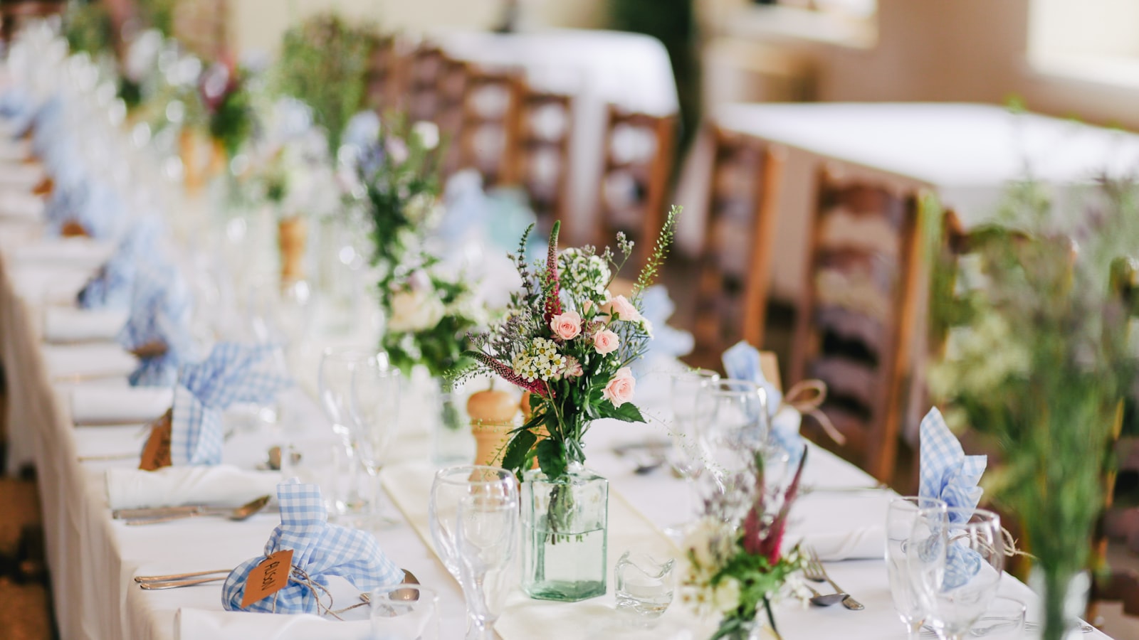 Una mesa larga de banquete puesta con manteles blancos, servilletas de cuadros azules y pequeños ramos silvestres en jarras de cristal, sin nadie sentado