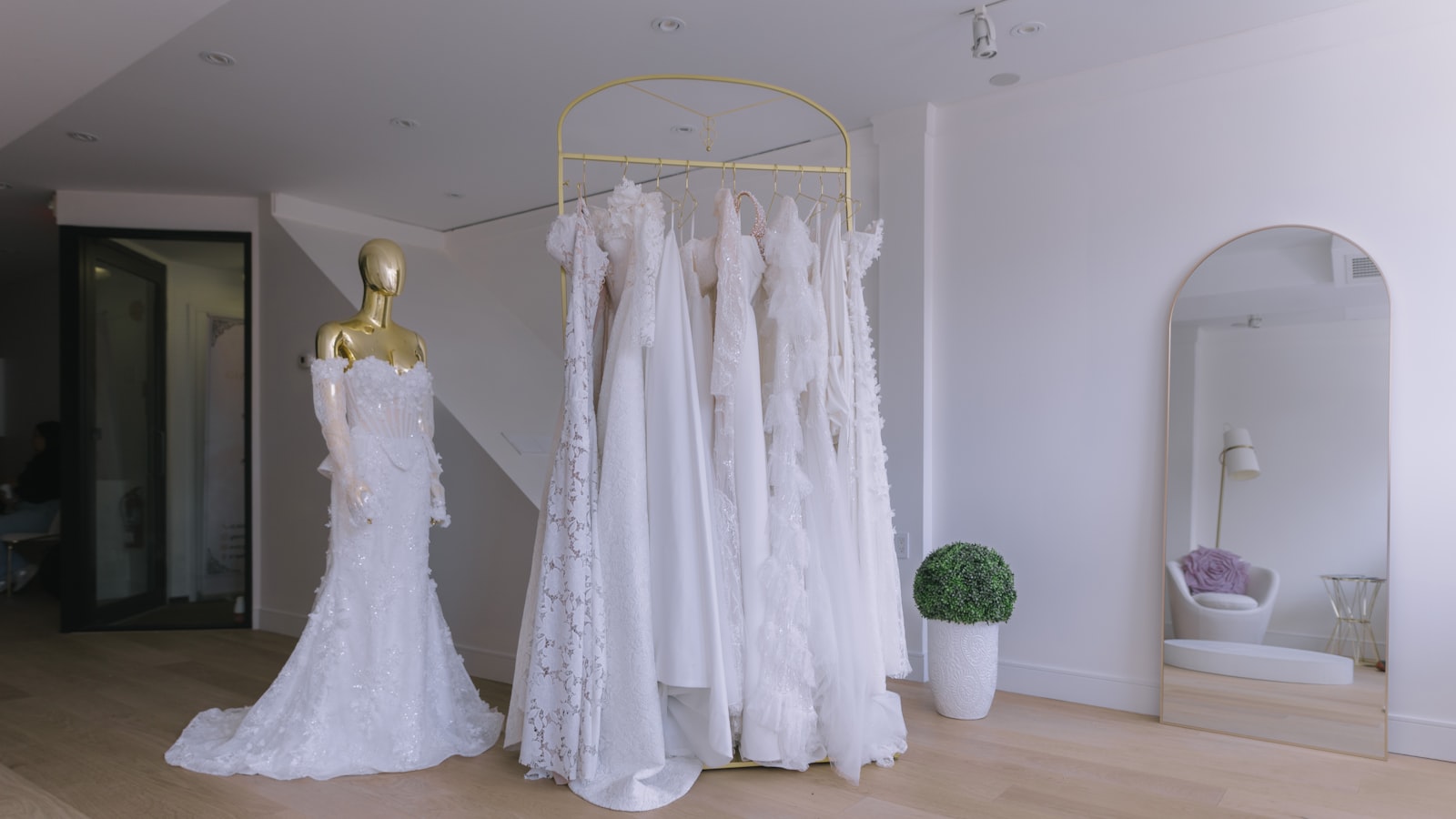 Vestidos de novia colgados en un perchero dorado en forma de arco, junto a un maniquí con un vestido de encaje, en una boutique de paredes blancas