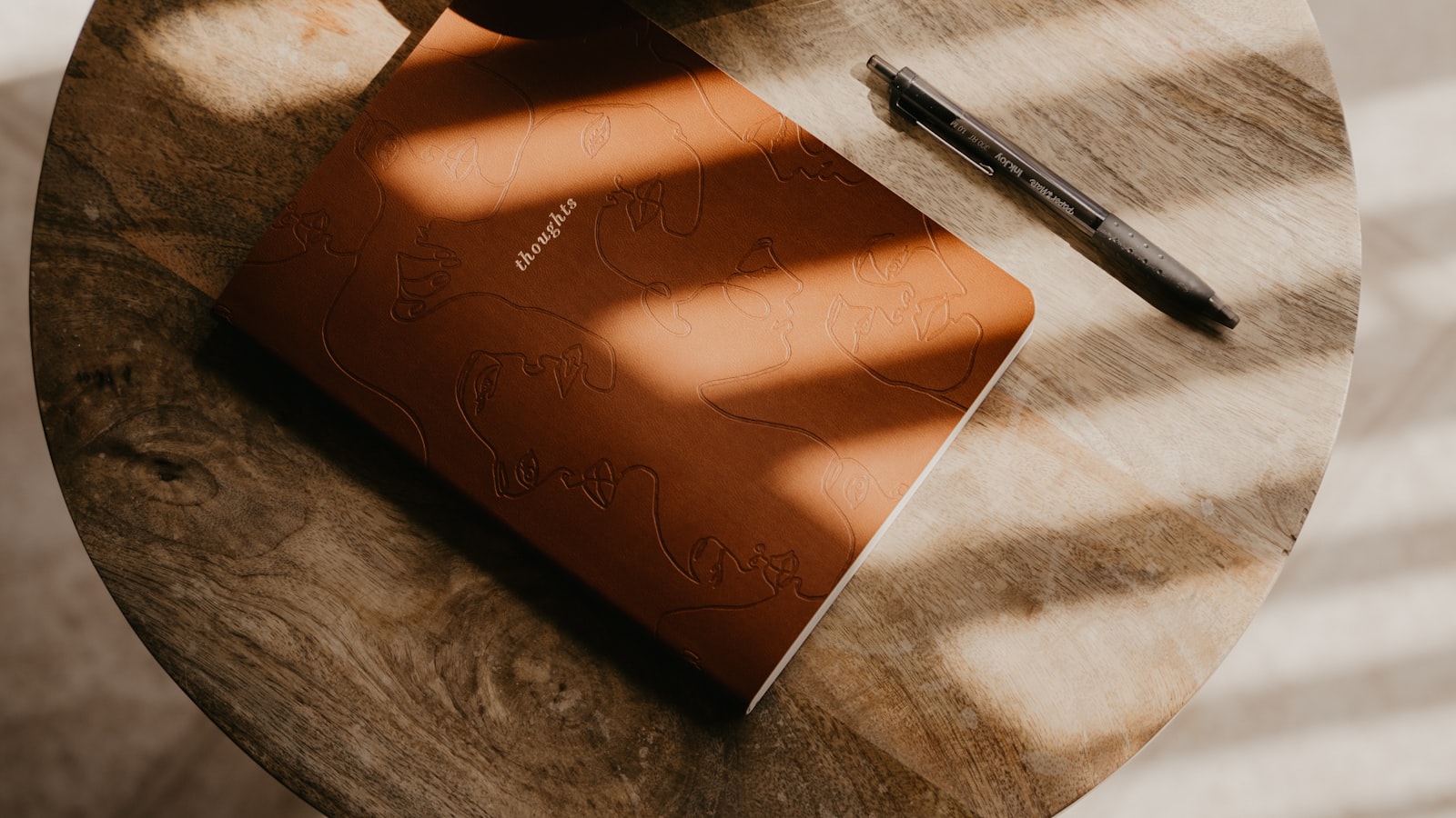 Una libreta de tapa marrón con la palabra "thoughts" y un bolígrafo sobre una mesa de madera, con luz de tarde entrando en líneas