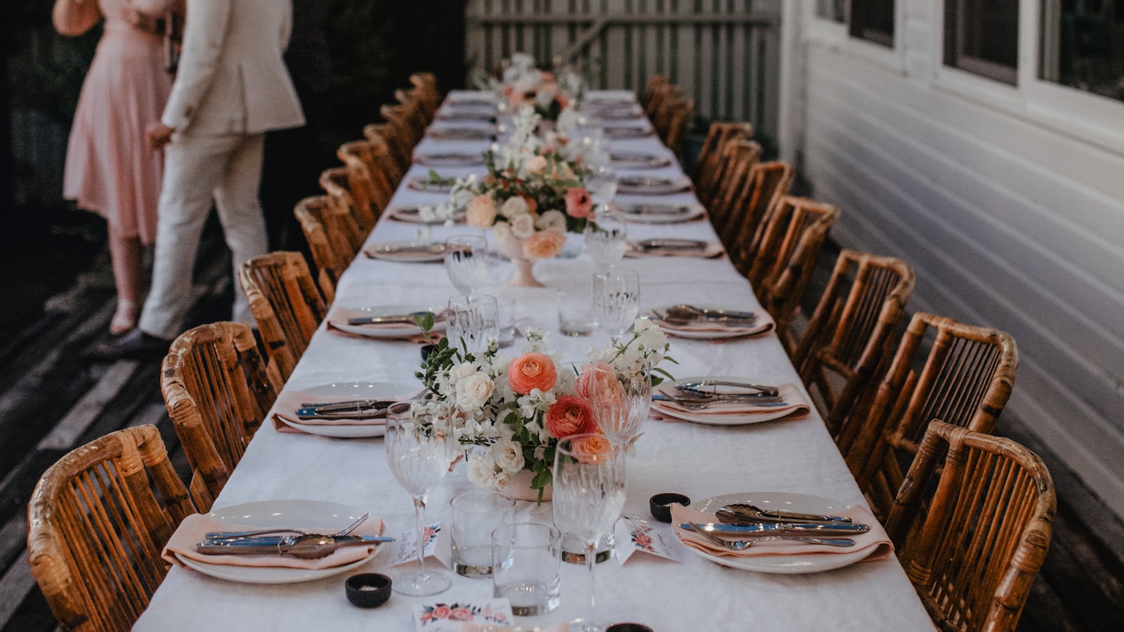 Una mesa larga puesta para una comida familiar al aire libre, con mantel blanco, centros de flores y sillas de mimbre a los dos lados