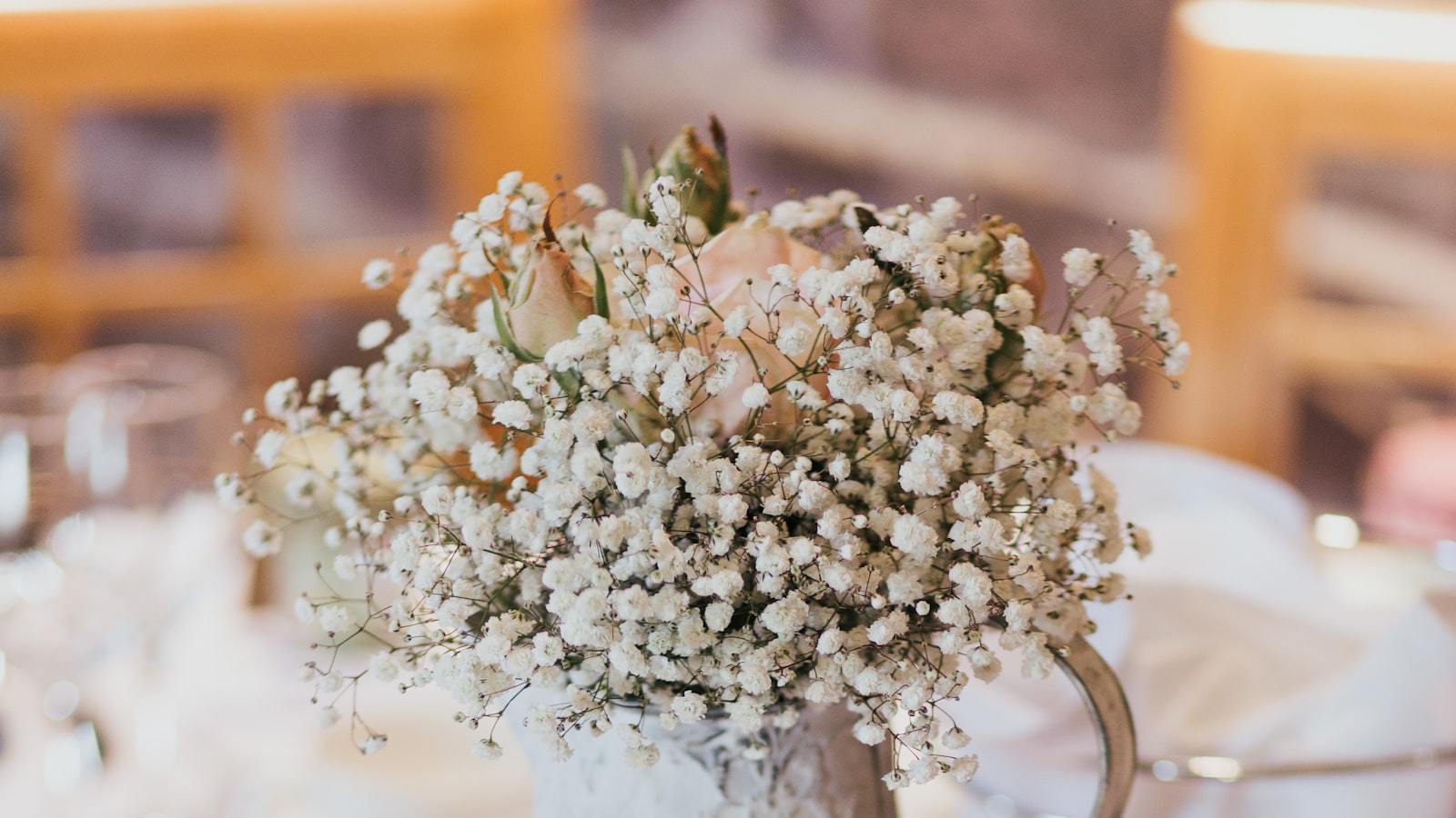 Un centro de mesa de flores blancas y una rosa pálida sobre un mantel claro, con copas y sillas desenfocadas al fondo de un salón de bodas