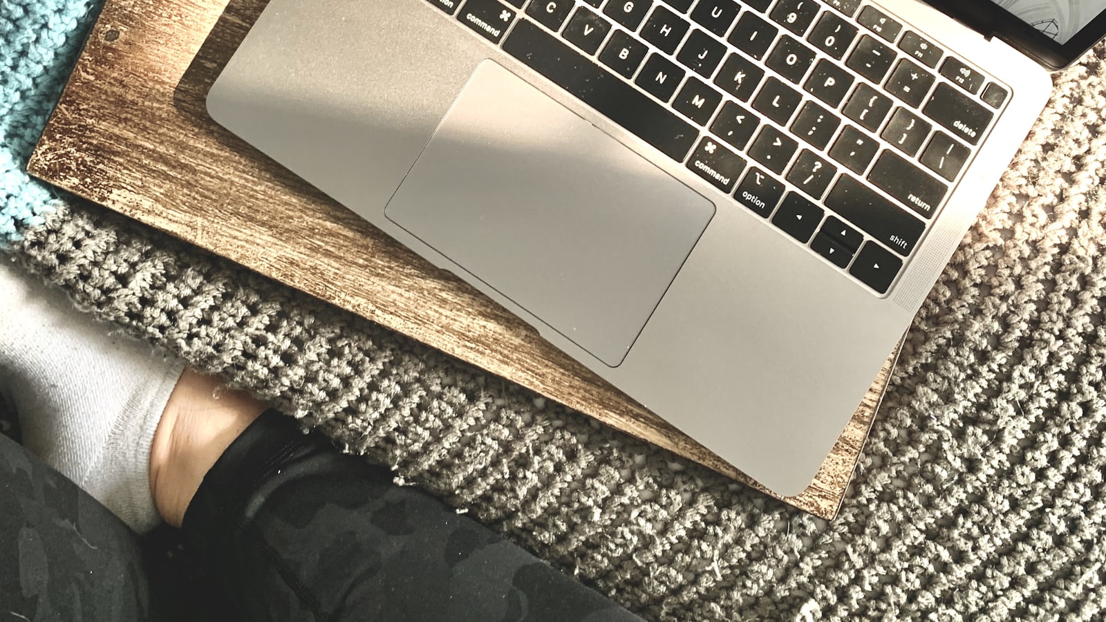 Un MacBook apoyado sobre una tabla de madera encima de un sofá, con un pie descalzo asomando bajo una manta de punto